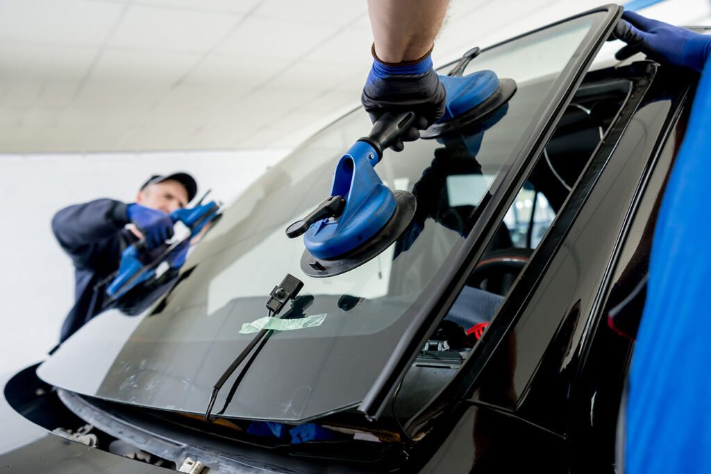 Two technicians wearing gloves are installing a car windshield. They use blue suction tools, focusing with precision and care in a bright workshop