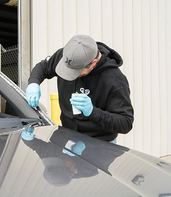 Man in a cap and hoodie repairs a car windshield, focused, wearing blue gloves, against a beige industrial building background